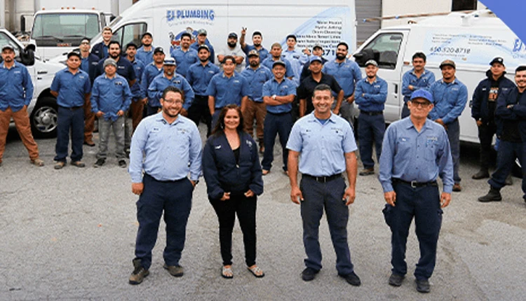 A group of 25 diverse plumbing team members in blue uniforms stand confidently in front of several company vans. The mood is professional and united.