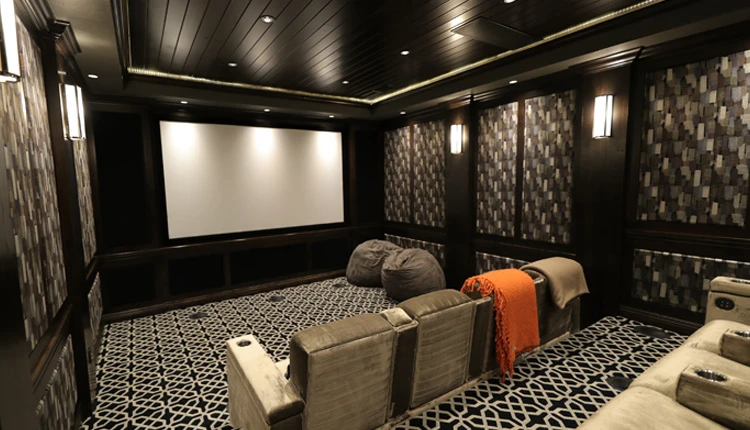 Luxurious home theater with a large screen, patterned carpet, plush gray recliners, bean bags, and a cozy orange throw, creating a warm, inviting ambiance.