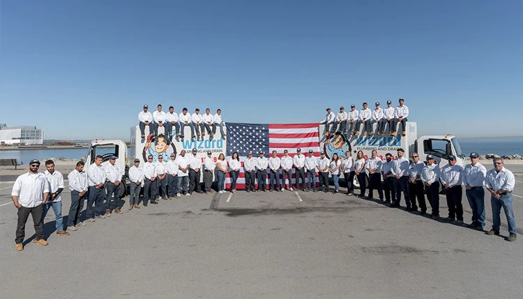 A group of people in uniform stands in front of two trucks, with an American flag between them. The sky is clear, and the mood is professional and unified.