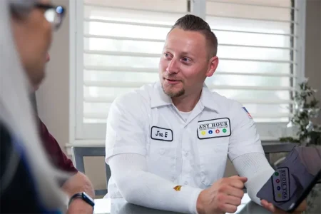 A man in a white work uniform sits at a table, smiling and gesturing while conversing with another person. A pleasant and professional atmosphere is conveyed.